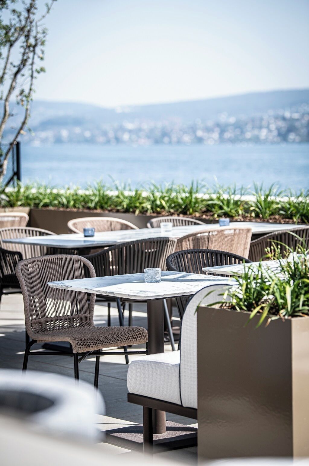 An outdoor seating area with wicker chairs and tables, overlooking a body of water and a distant cityscape.