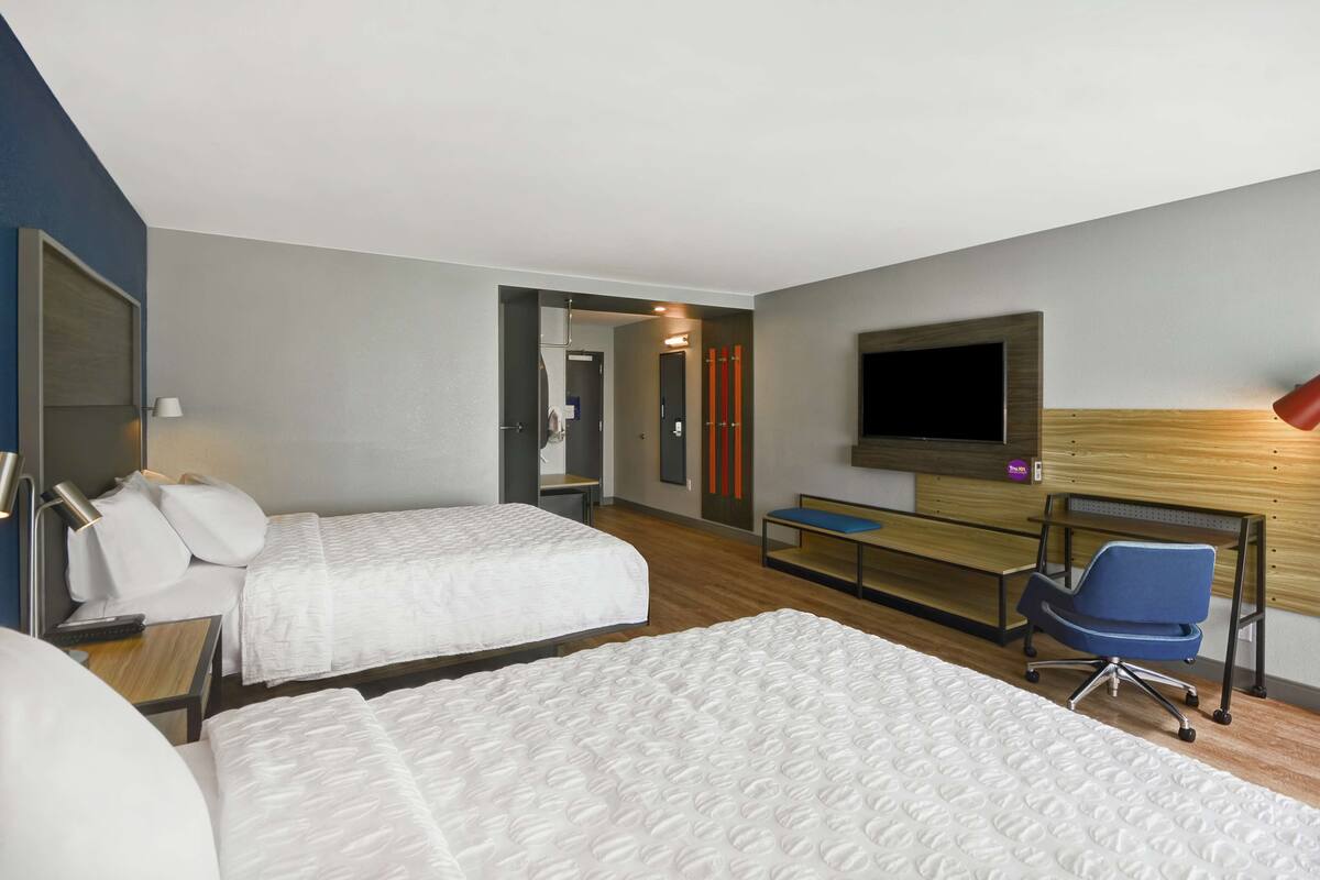 Habitació d'hotel amb dos llits, un escriptori, una cadira, un televisor i un panell de capçaló de fusta.