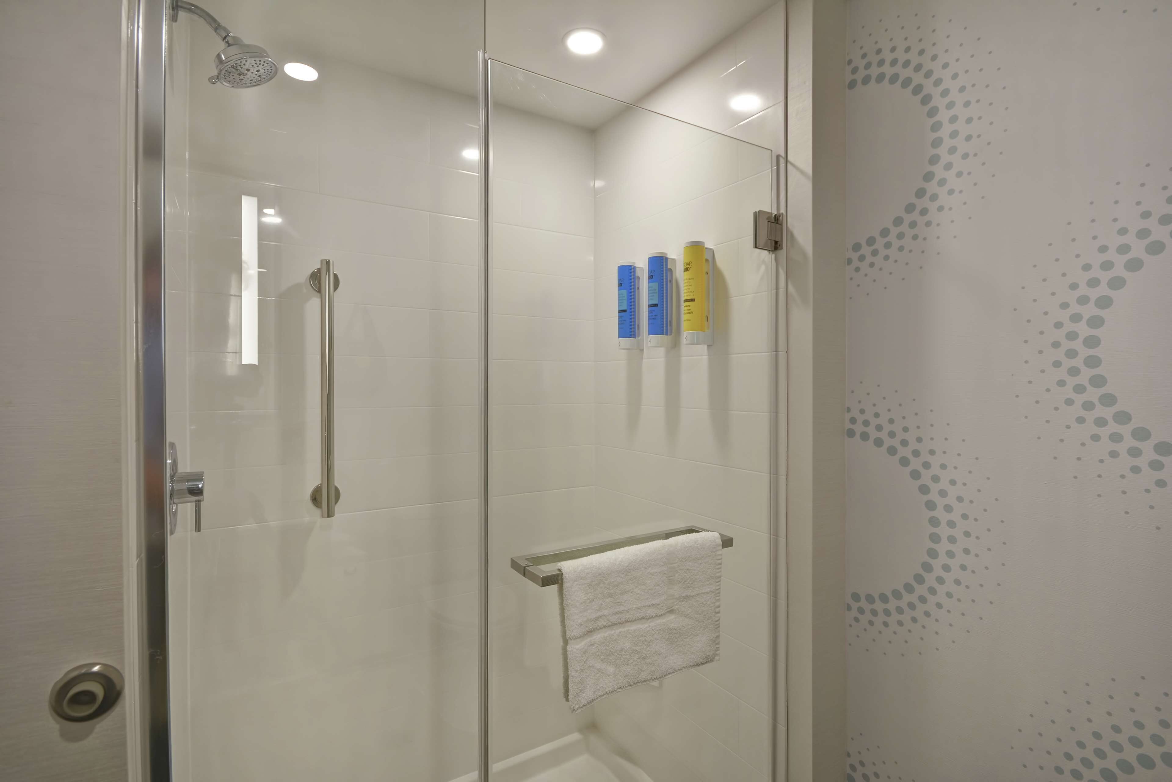 Um chuveiro com divisória de vidro, um chuveiro manual, um porta-toalhas com uma toalha branca e duas garrafas de shampoo na parede.