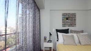 A bedroom with a bed, a nightstand, a lamp, a wall art piece, and a window with curtains. - Edem Beach Apartment Mountain View (Palaio Faliro)