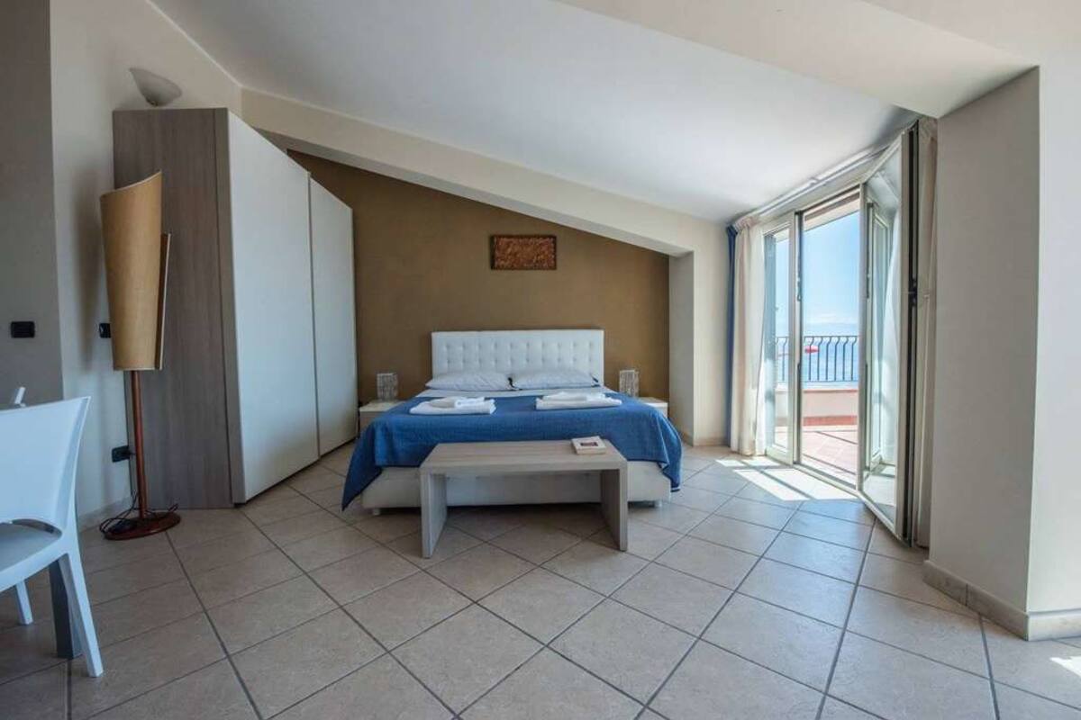 A bedroom with a bed, a bedside table, a chair, a floor lamp, and a view of the outside.