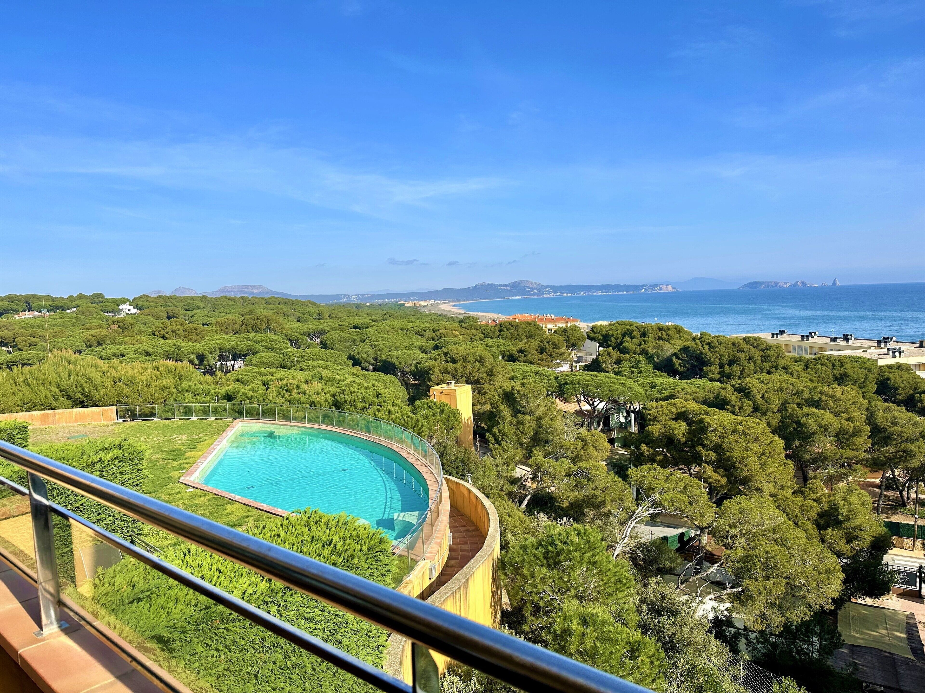 Piscina com borda curva, rodeada por vegetação exuberante e vista clara para o mar e montanhas ao longe.