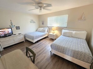 A bedroom with two beds, a desk with a television, a chair, a ceiling fan, and a window with blinds.