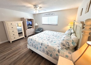 Um quarto com cama, cômoda, televisão, cadeira e ventilador de teto.