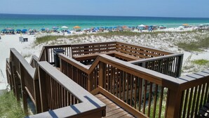 Eine Holzterrasse mit Geländern, die auf einen Strand mit Sonnenschirmen und Menschen blickt.