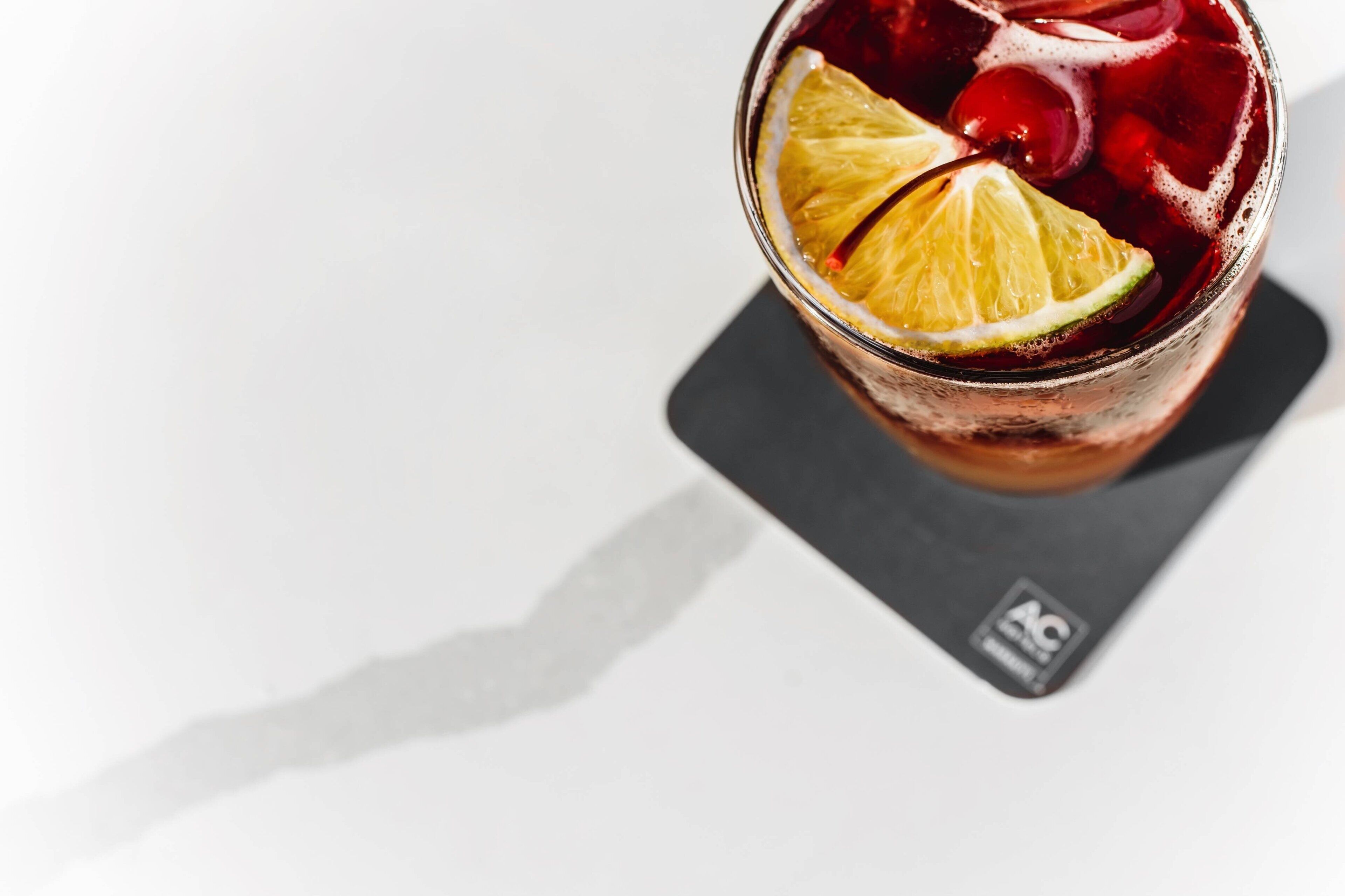 A glass of beverage with a slice of lime and a cherry, placed on a coaster with a logo.