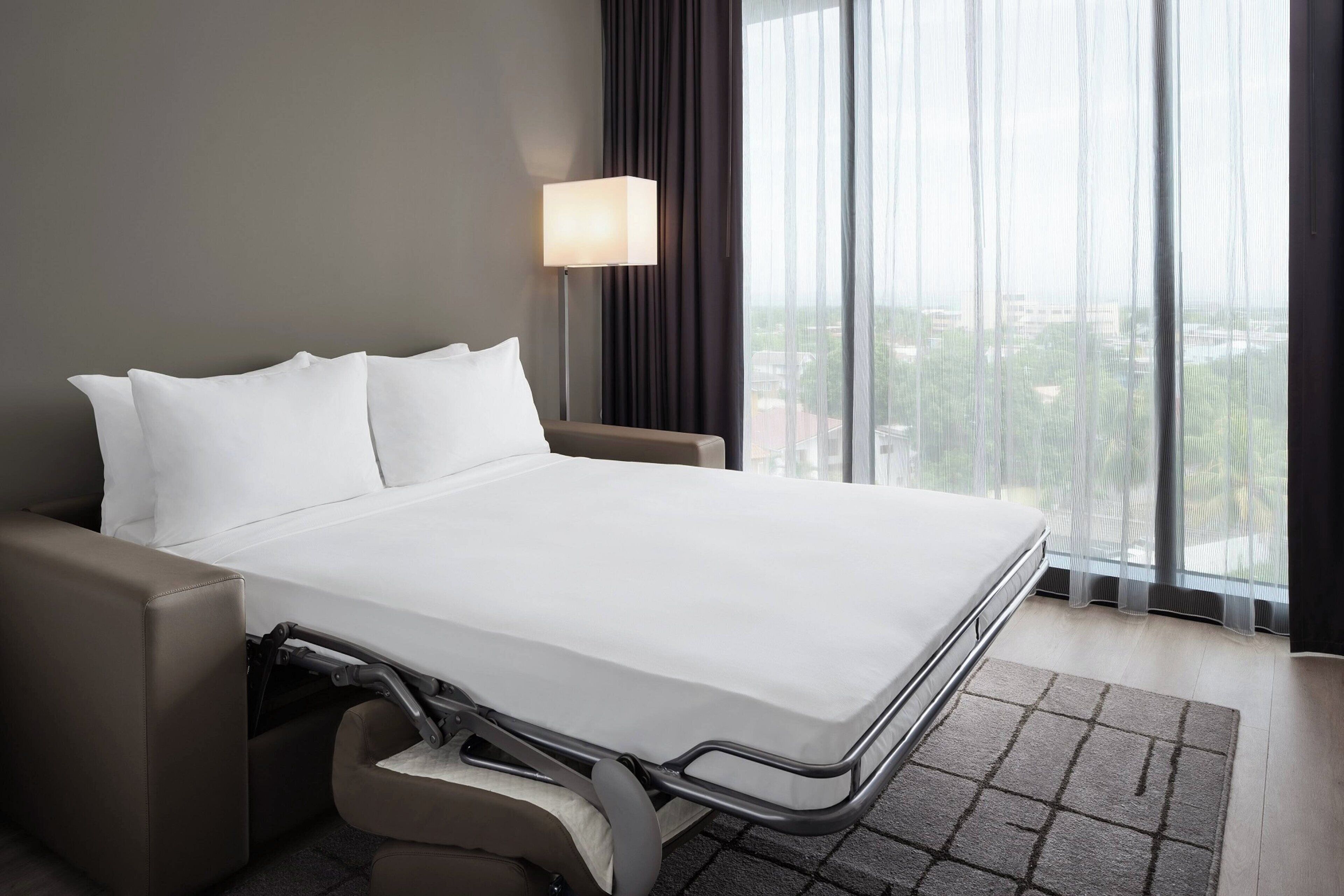 A hotel room with a bed, a desk, a chair, a lamp, and a view of the city through large windows.