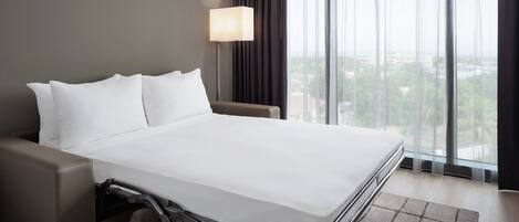 A hotel room with a bed, a desk, a chair, a lamp, and a view of the city through large windows.