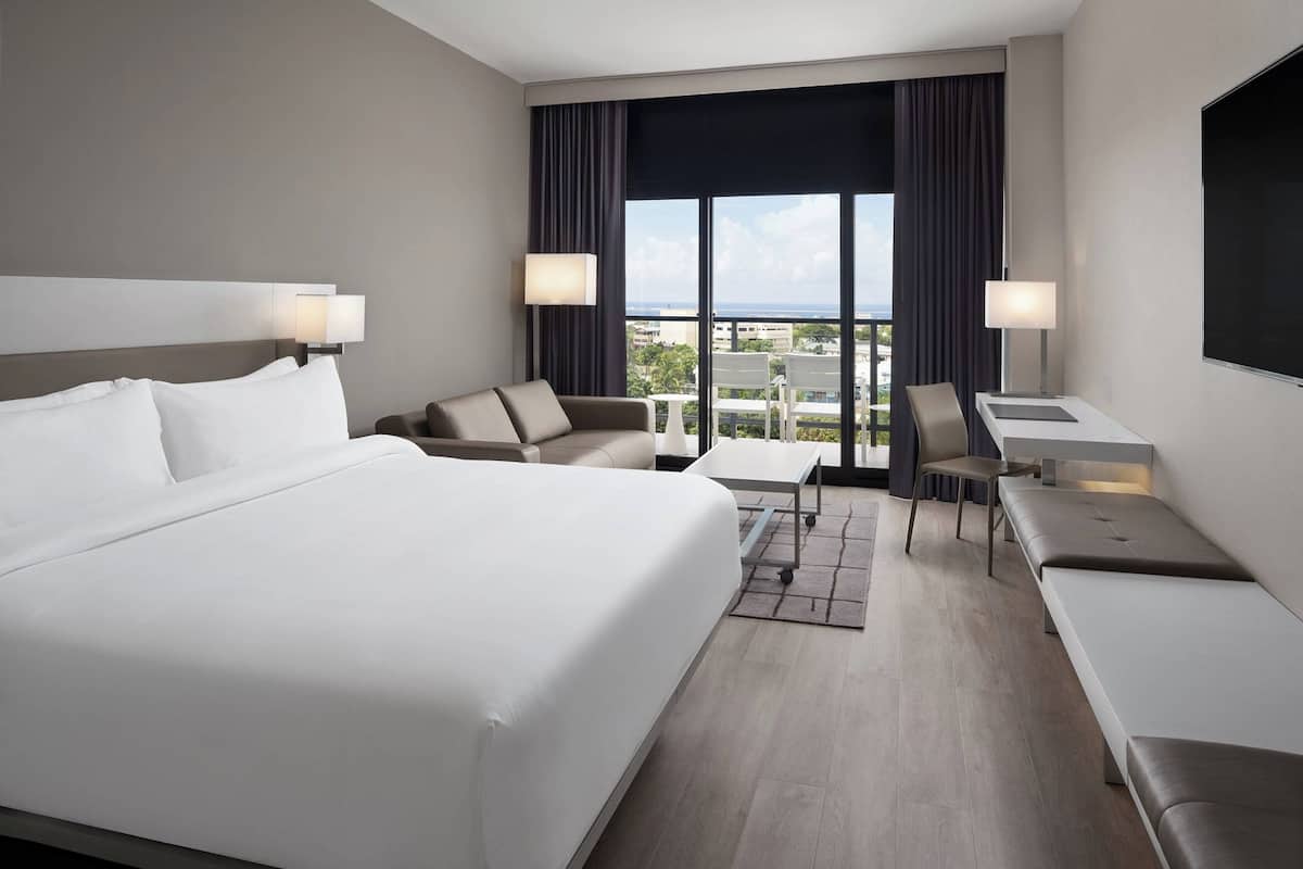 Une chambre d’hôtel avec un grand lit, un canapé, un bureau et une chaise. Elle comporte deux fenêtres avec des rideaux, et on peut admirer la vue sur la ville à travers ces fenêtres.