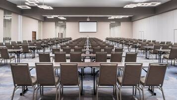 Un amplio salón de conferencias con filas de mesas y sillas, una pantalla de proyector y suelo alfombrado.