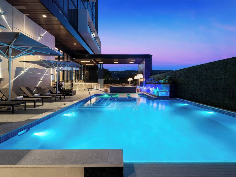 A modern outdoor pool area with lounge chairs and umbrellas, a bar, and a view of the city at dusk.