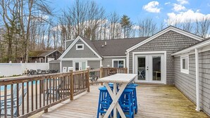 A deck with a table and blue stools, surrounded by a wooden railing, with a house in the background.