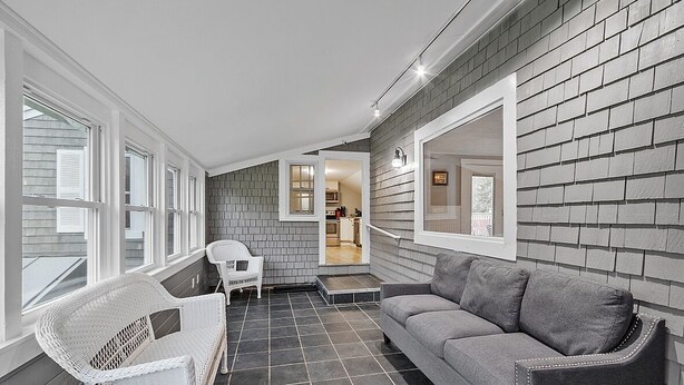 A compact living space with a grey brick wall, a grey sofa, a white wicker chair, and a small table. A kitchen is visible through an open doorway.