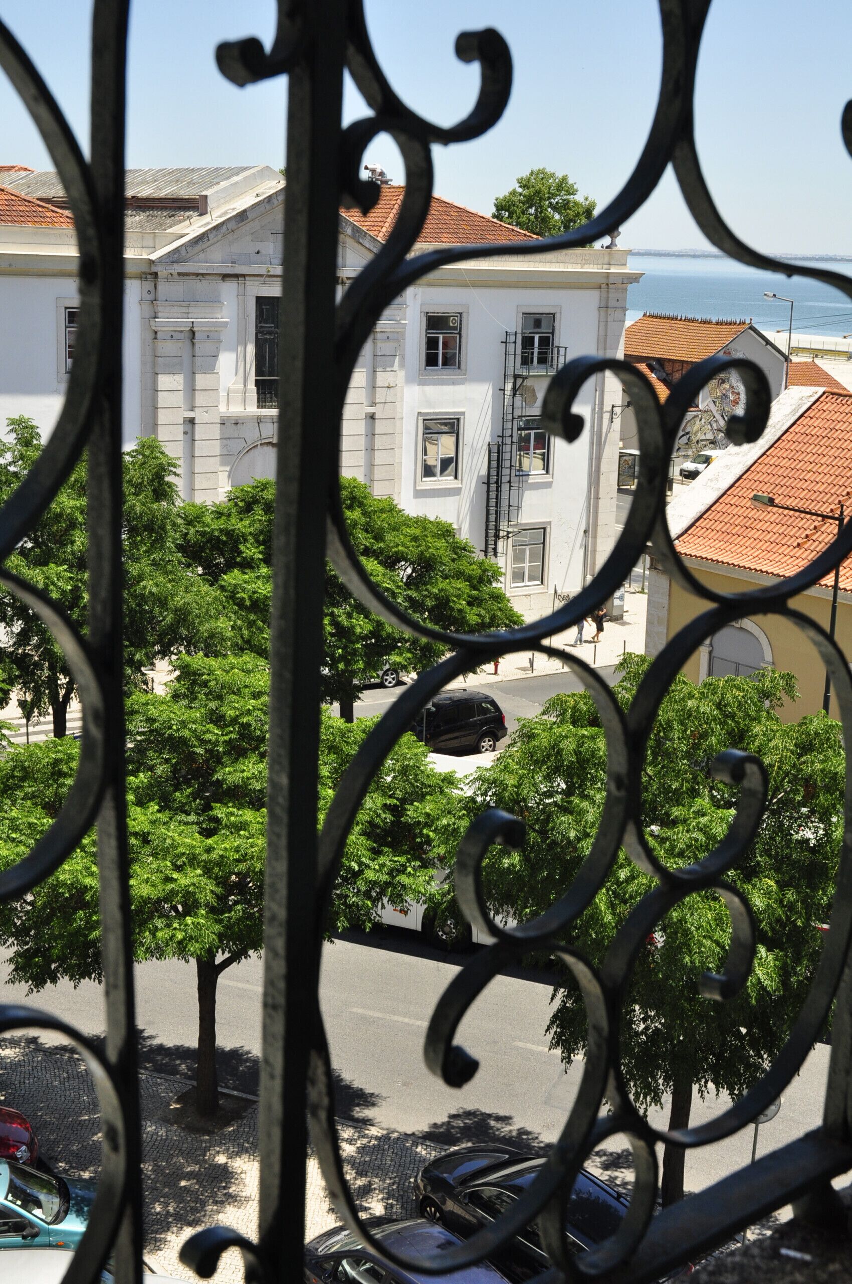 A view through ornate metalwork, featuring a street with parked cars, trees, and buildings with red-tiled roofs.