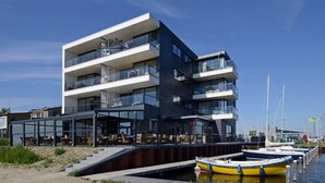 Un immeuble moderne à plusieurs étages, doté de balcons, d’une façade en verre et situé en bord de mer, avec des bateaux amarrés.