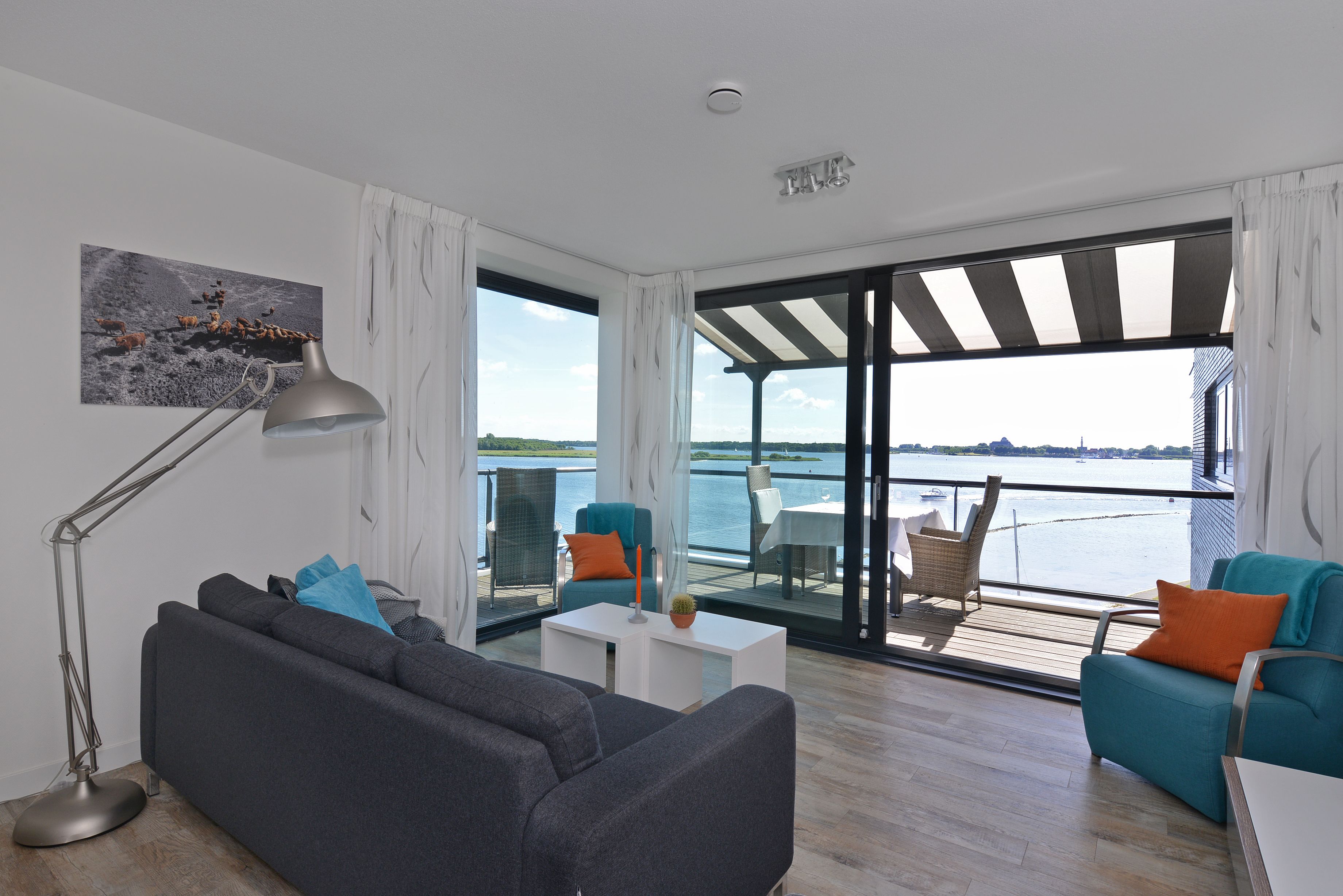 A modern living room with a sofa, a coffee table, and two chairs. There is a large window with a view of the water and a balcony.