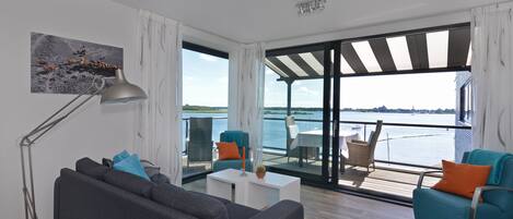 A modern living room with a sofa, a coffee table, and two chairs. There is a large window with a view of the water and a balcony.