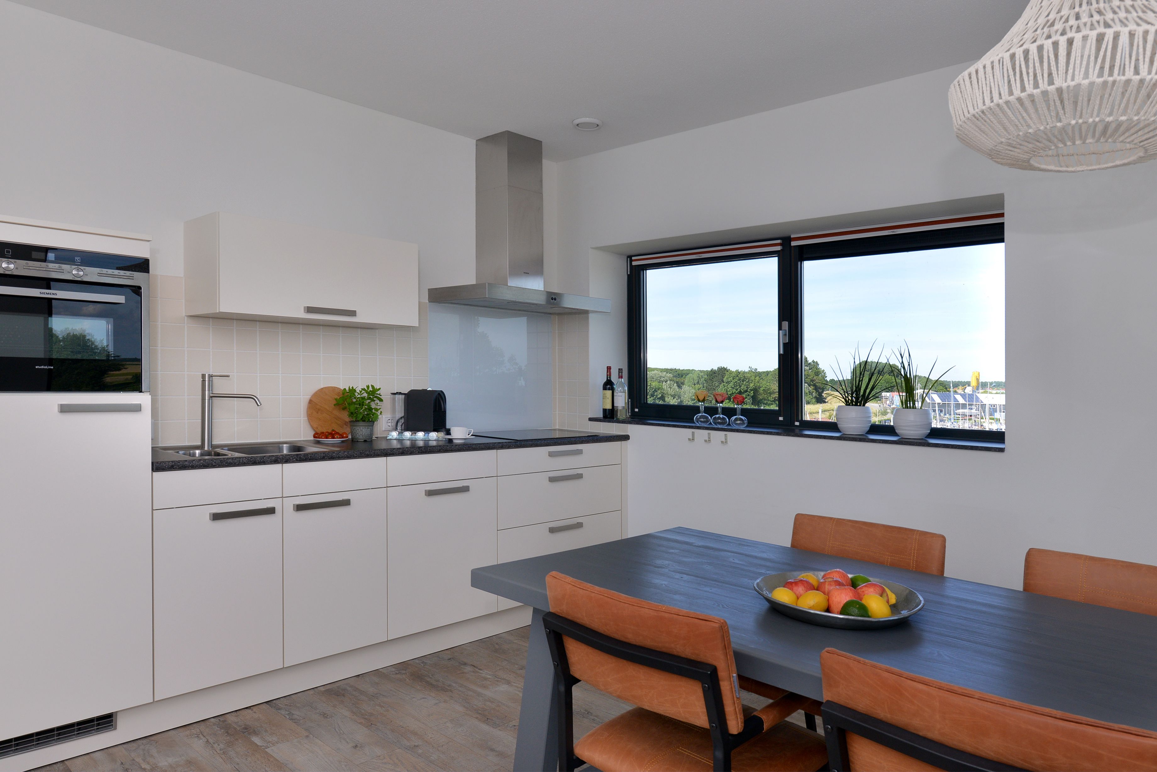 Une cuisine moderne avec des meubles blancs, un four encastré et une fenêtre donnant sur un paysage verdoyant.