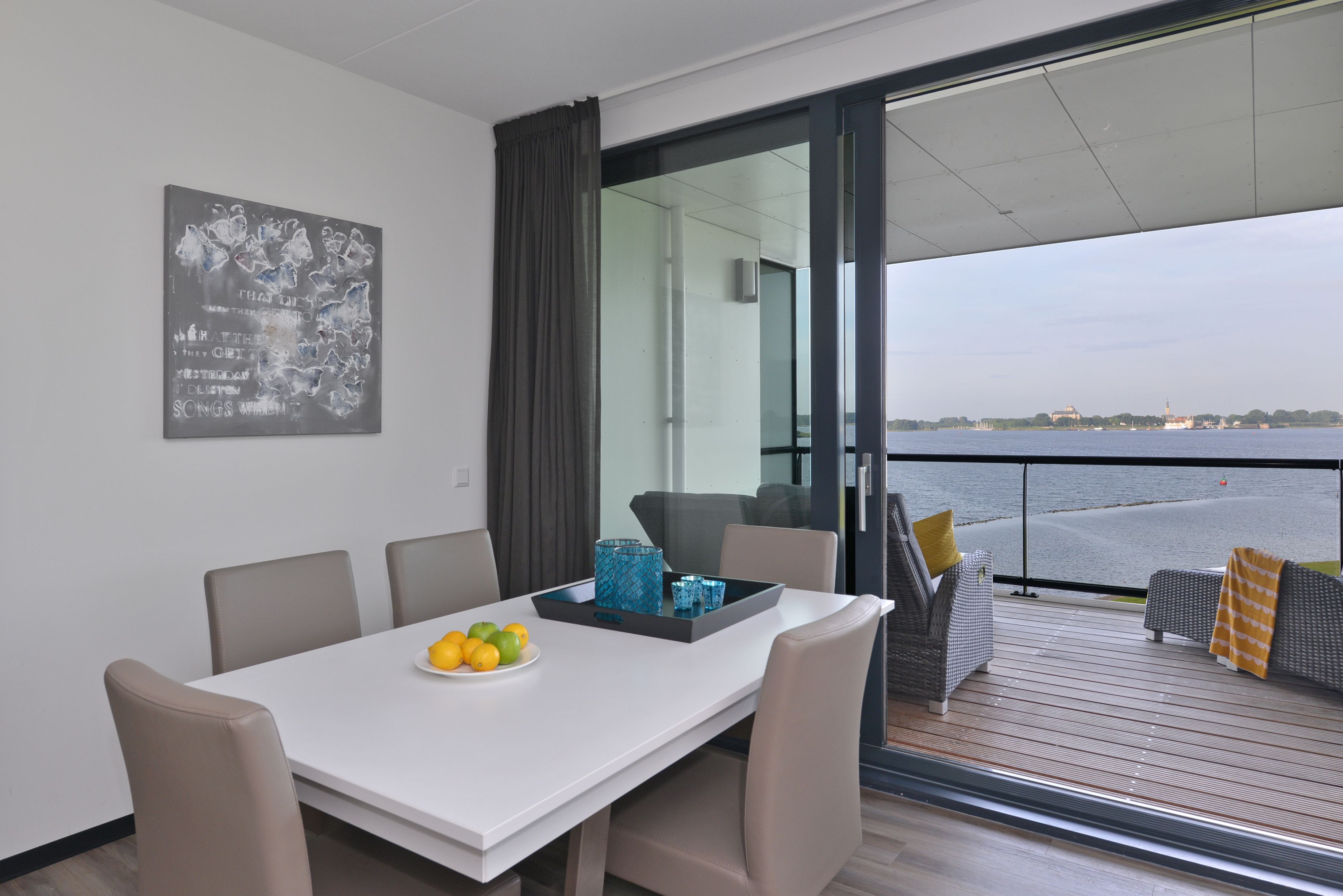 A modern dining area with a white table, beige chairs, and a glass partition. A tray with lemons and a blue decorative item is on the table. Outside, there is a balcony with grey wicker furniture and a view of the sea.