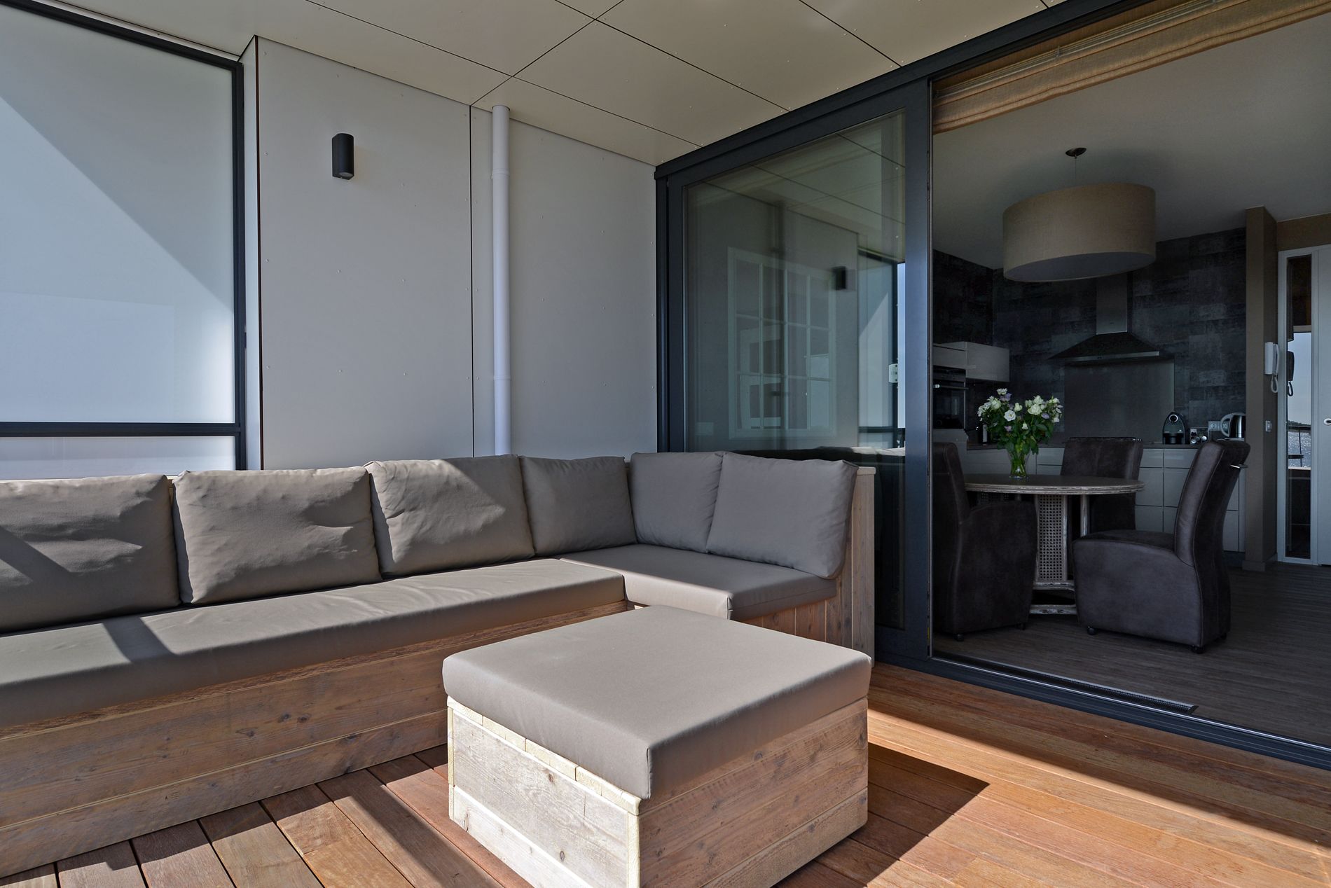 A modern living room with a sectional sofa and ottoman, a glass door leading to a dining area, and a kitchen visible through the glass.