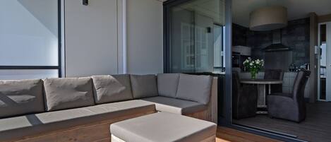 A modern living room with a sectional sofa and ottoman, a glass door leading to a dining area, and a kitchen visible through the glass.