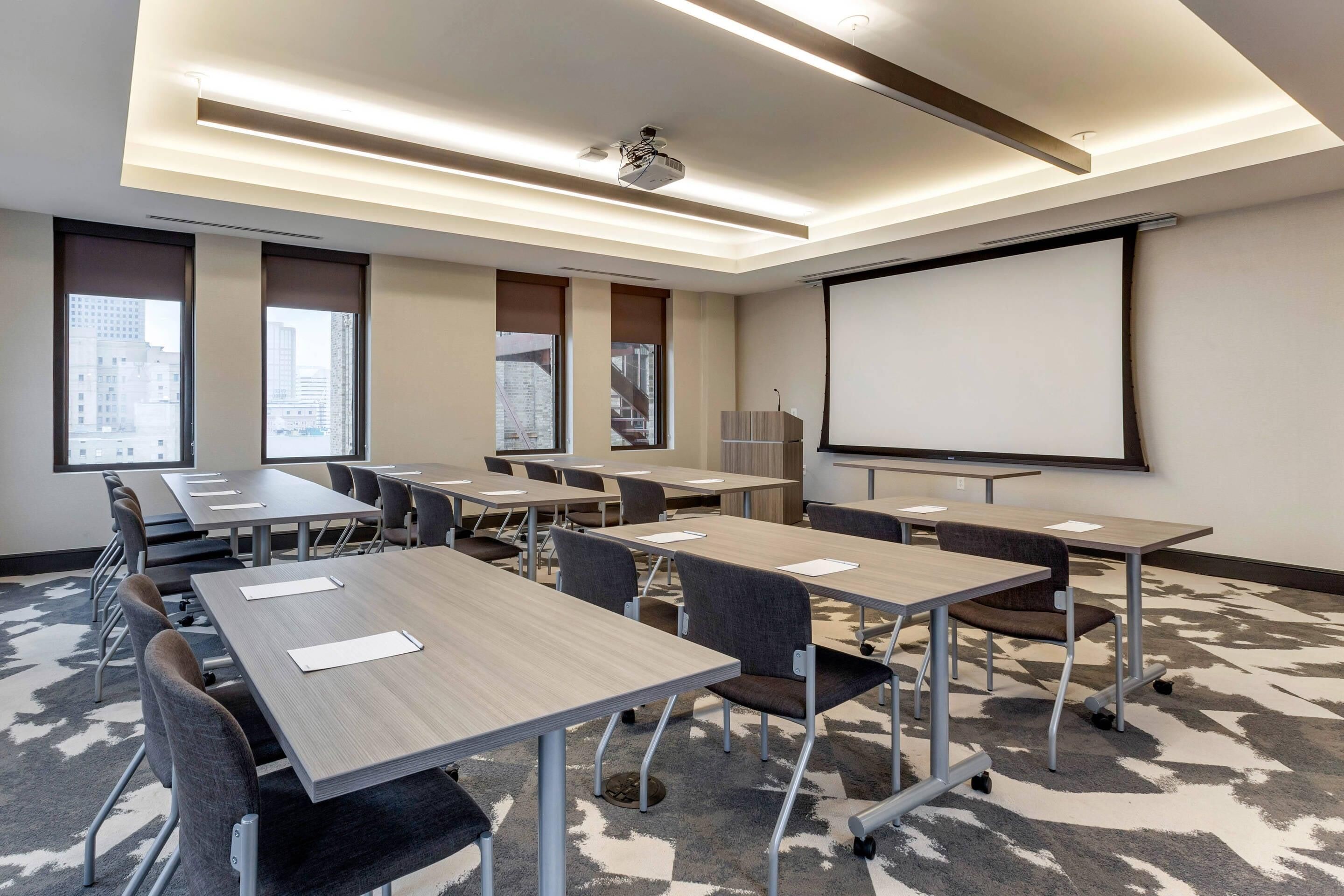 A conference room with multiple tables and chairs, a projector screen, and large windows with city views.