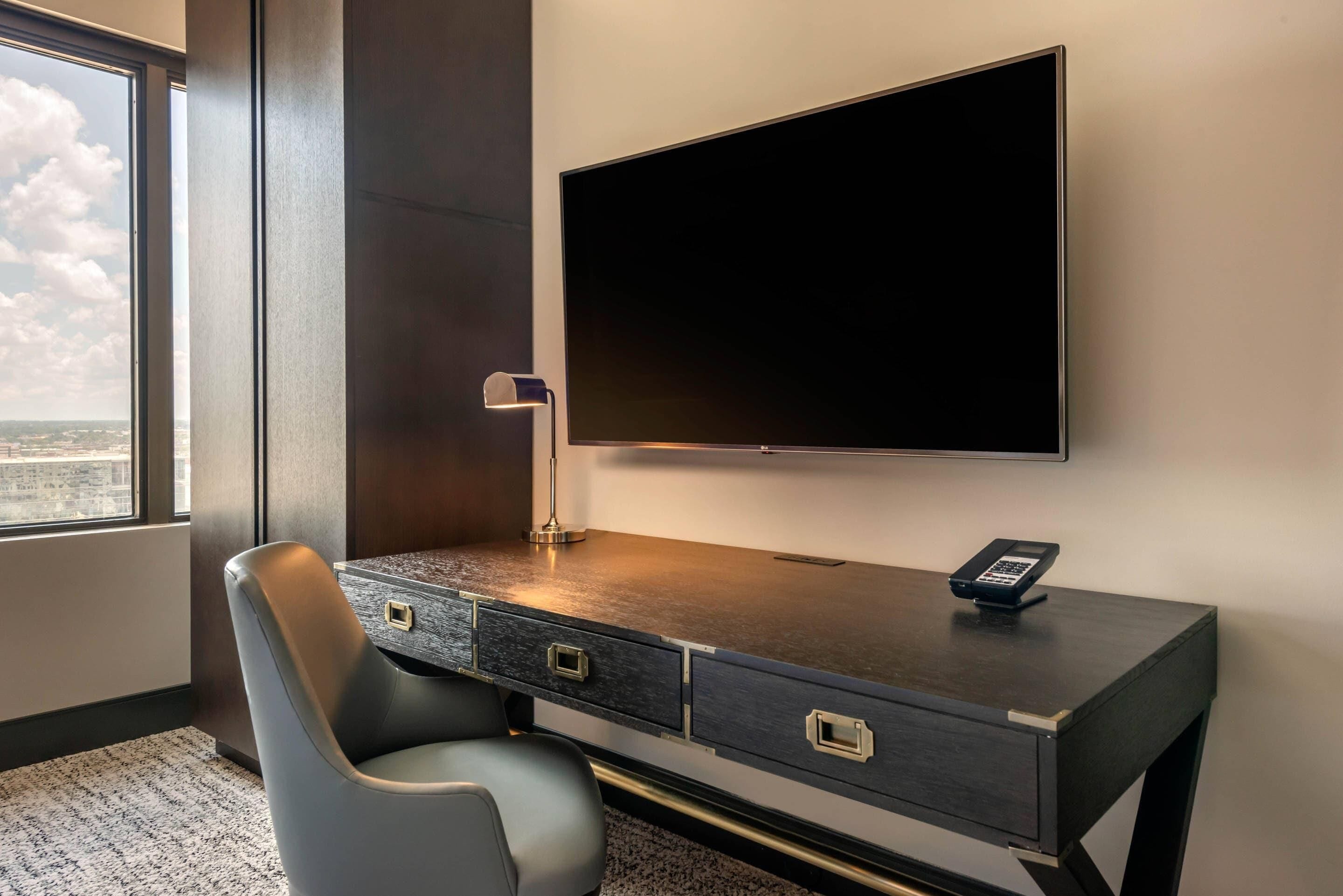 Ett modernt hotellrum med en stor platt-TV som är monterad på väggen, ett skrivbord med en lampa och en stol.