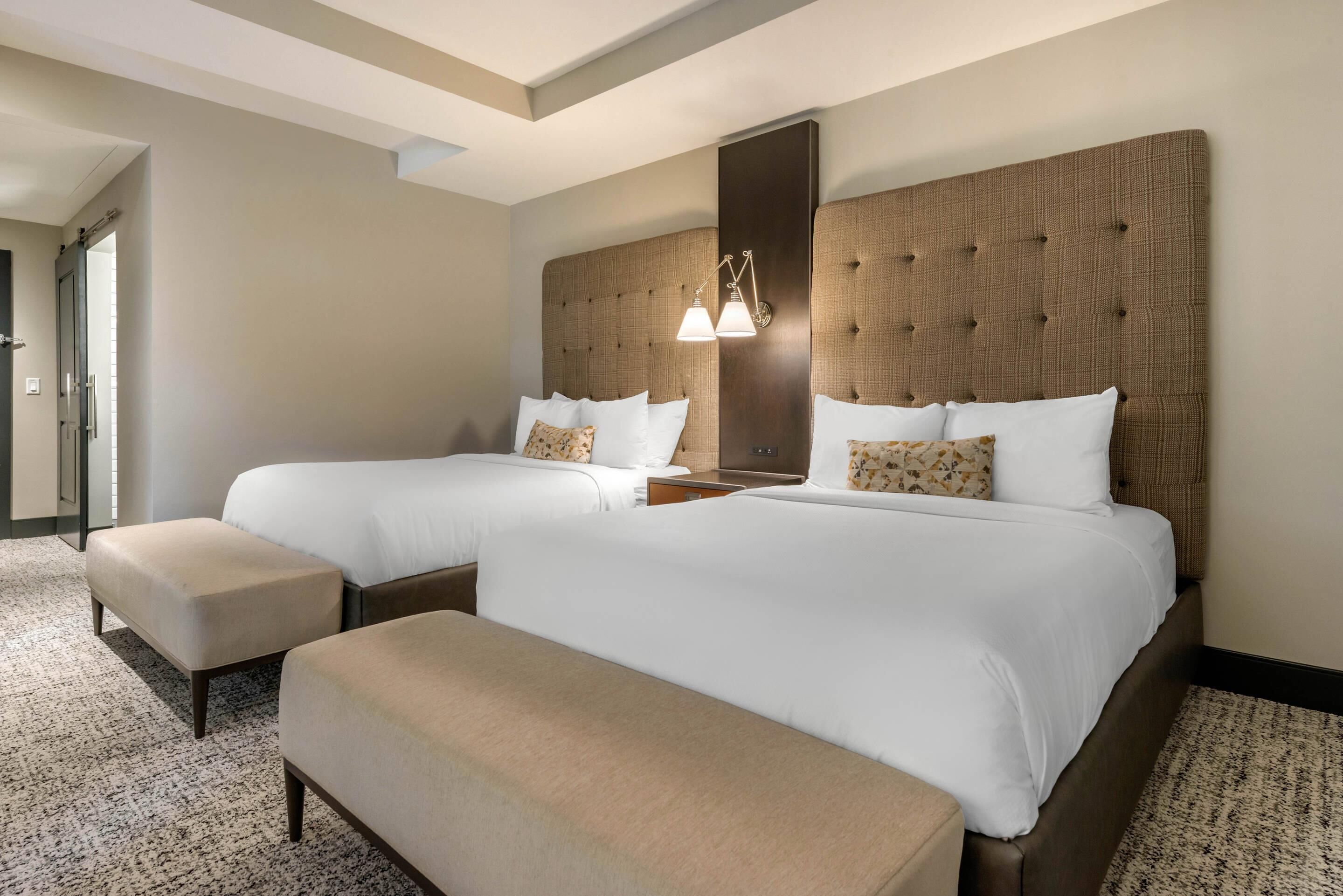 A hotel room with two beds, a large window, a headboard with a light fixture, and a carpeted floor.