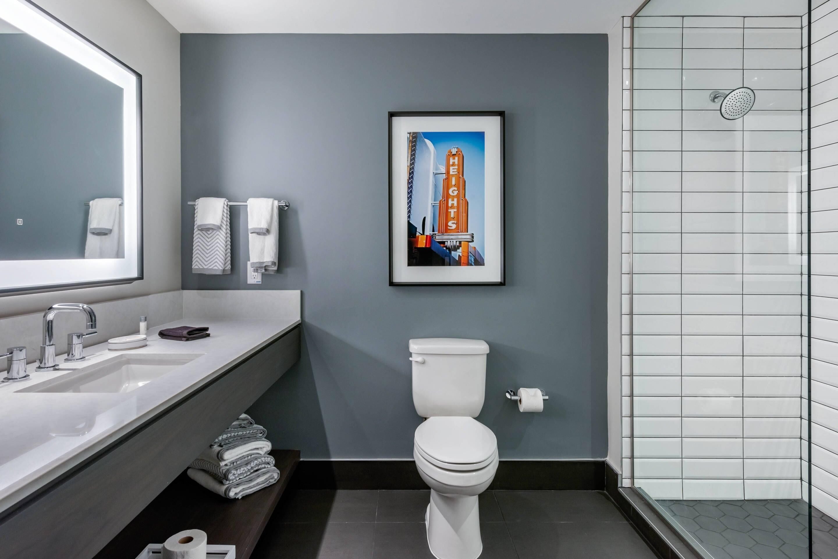 A modern bathroom with a large mirror, a sink, a toilet, and a shower. There is a framed picture of a building on the wall.