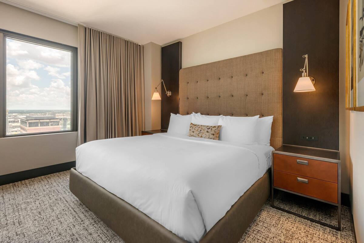 Ein Hotelzimmer mit einem großen Bett, einem Nachttisch mit Lampe und Blick auf die Stadt durch ein großes Fenster.