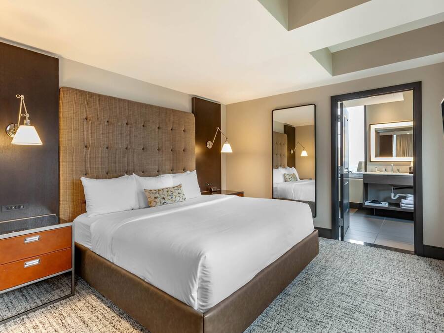 A modern hotel room with a large bed, a nightstand with drawers, a wall-mounted lamp, a large mirror, and a view into the bathroom.
