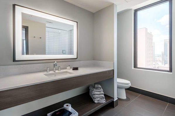 A modern bathroom with a large mirror, a sink, and a toilet. There is a window providing a view of buildings outside.