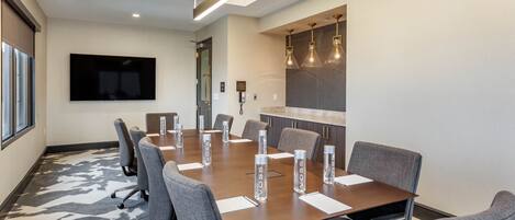 Un salón de conferencias con una mesa larga, sillas, un televisor de pantalla plana y una zona de cocina con lámparas colgantes.