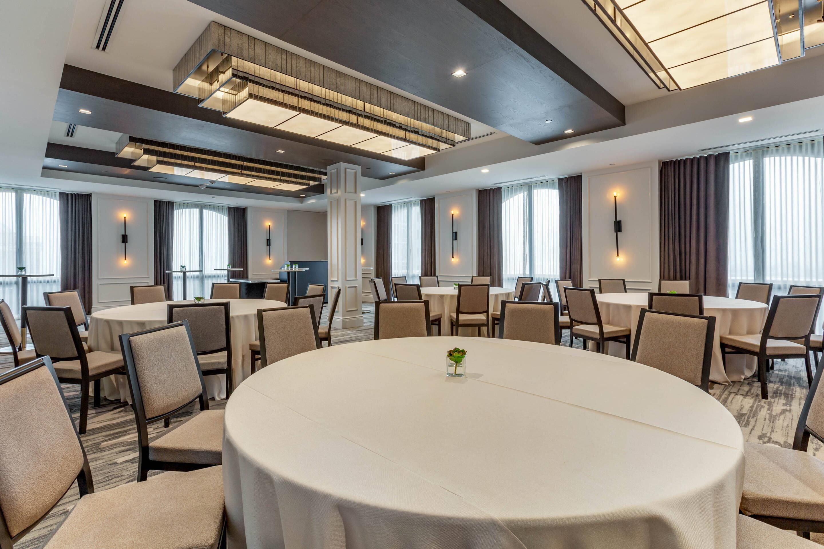 A conference room with round tables and chairs, large windows with curtains, and modern lighting fixtures.