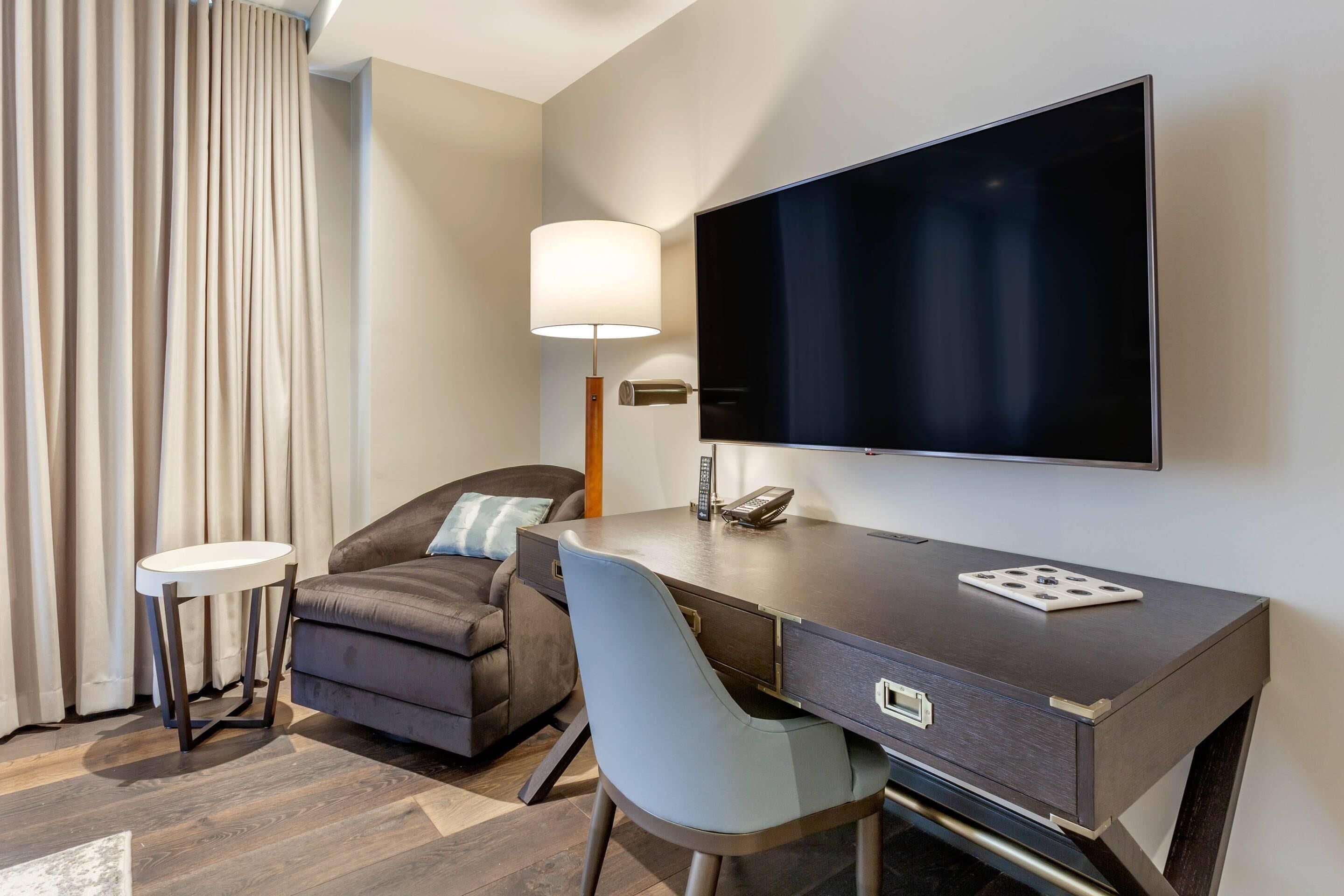A hotel room with a dark wood desk, a flat-screen TV, a sofa, a lamp, and a small side table.