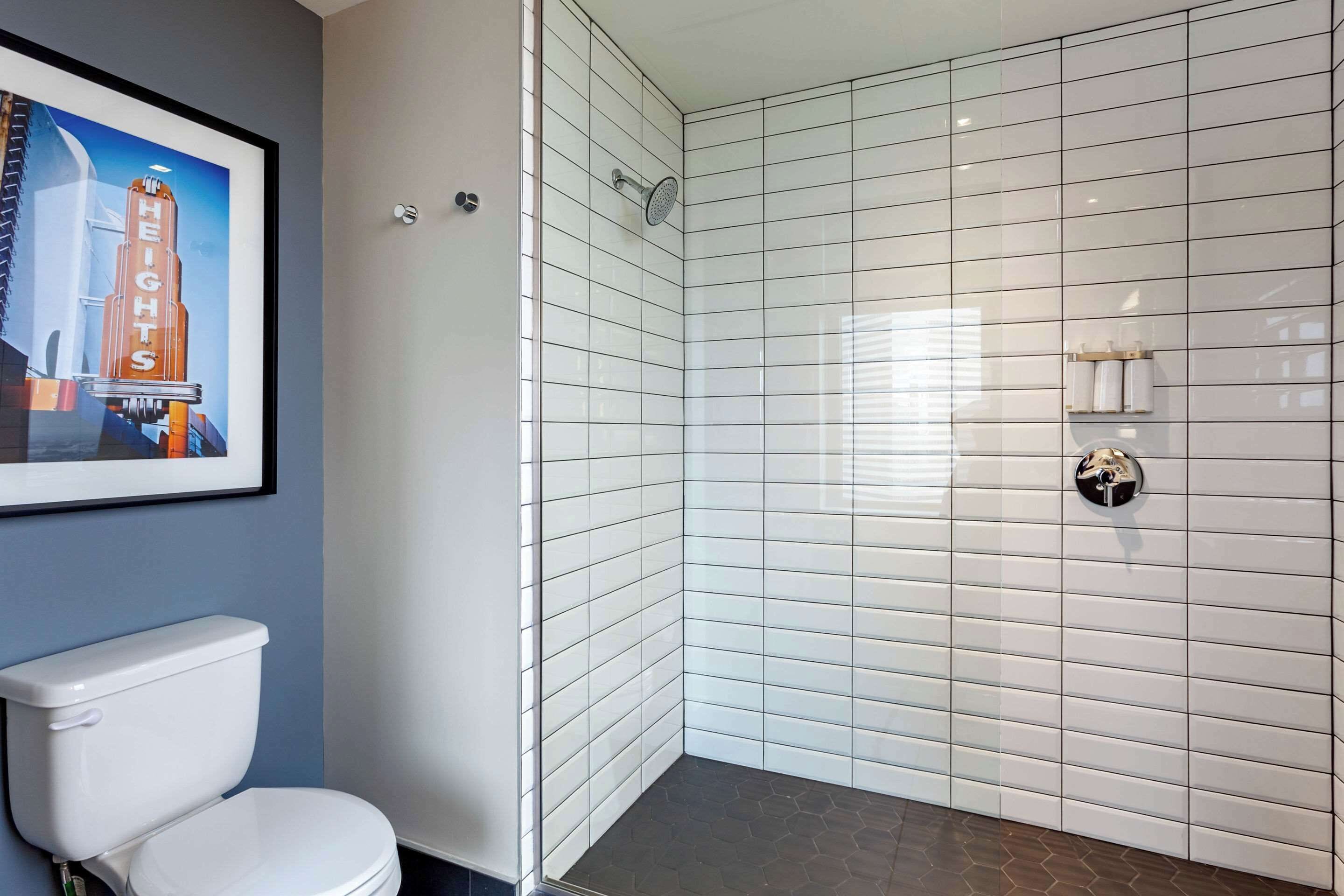 Baño con inodoro blanco, cabina de ducha de vidrio y pared de azulejos con un cuadro enmarcado.
