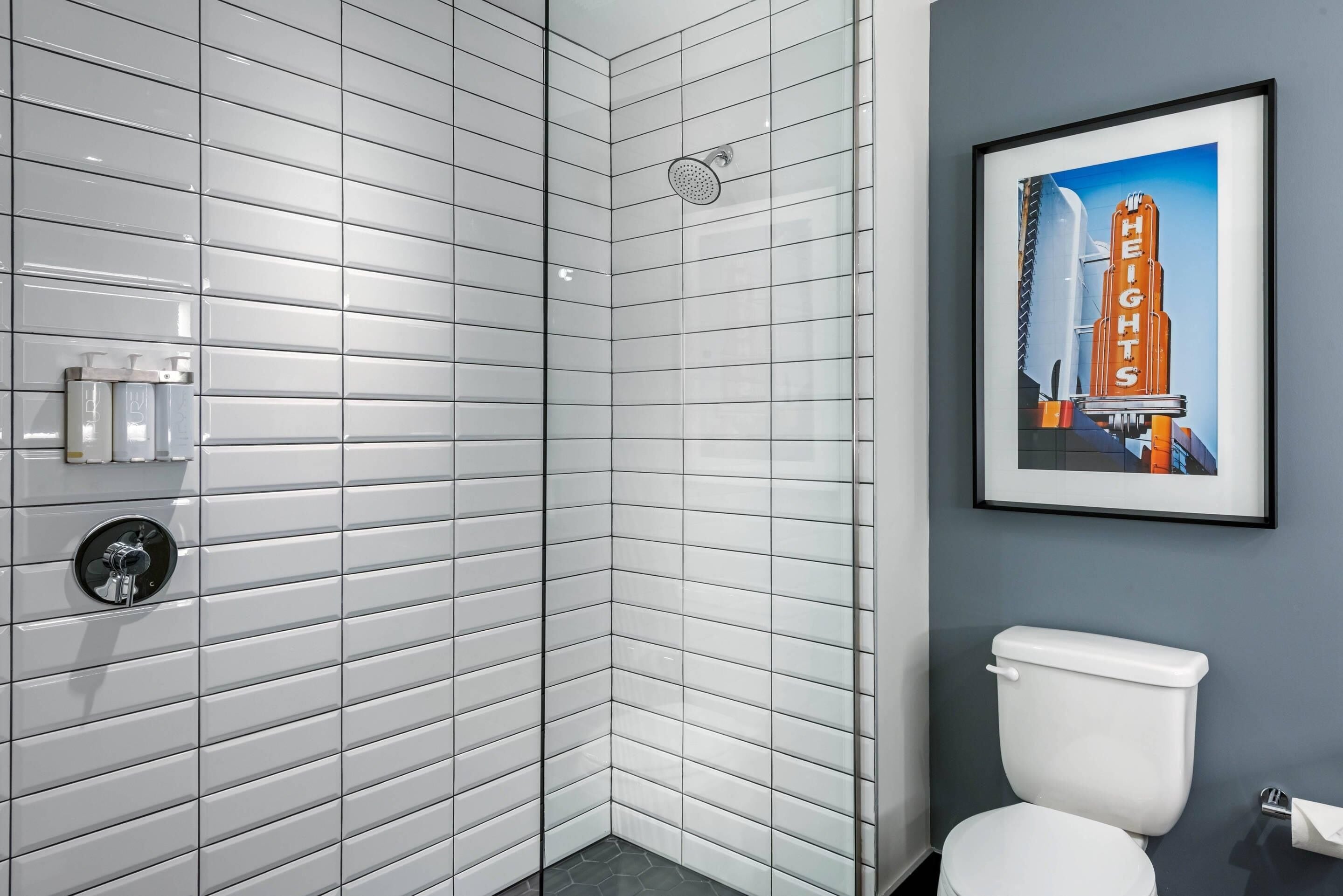A bathroom with white subway tile walls, a white toilet, a shower with a glass door, and a framed picture of a building on the wall.