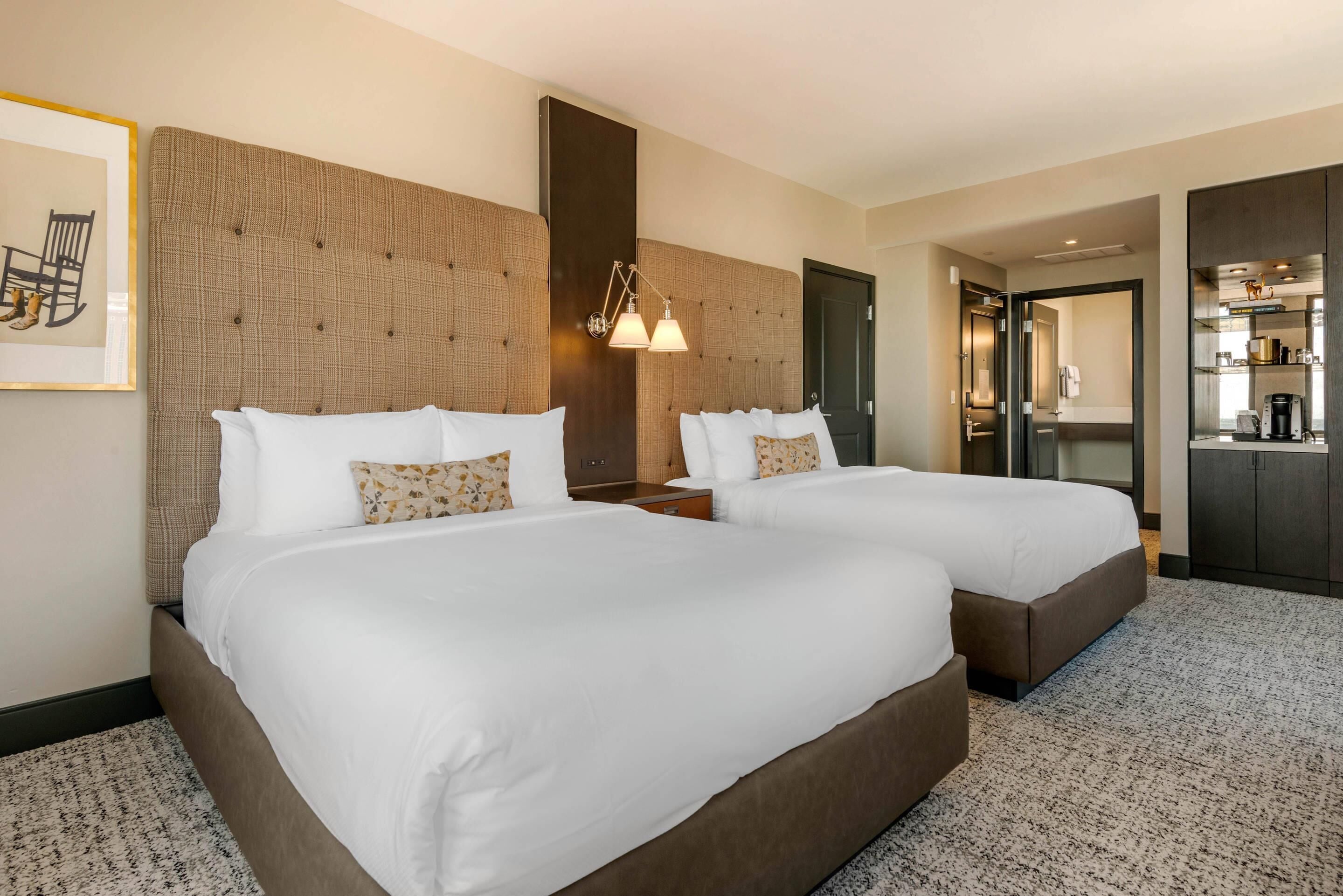 Habitación de hotel con dos camas, un cabecero grande, una lámpara de pared e imagen enmarcada.