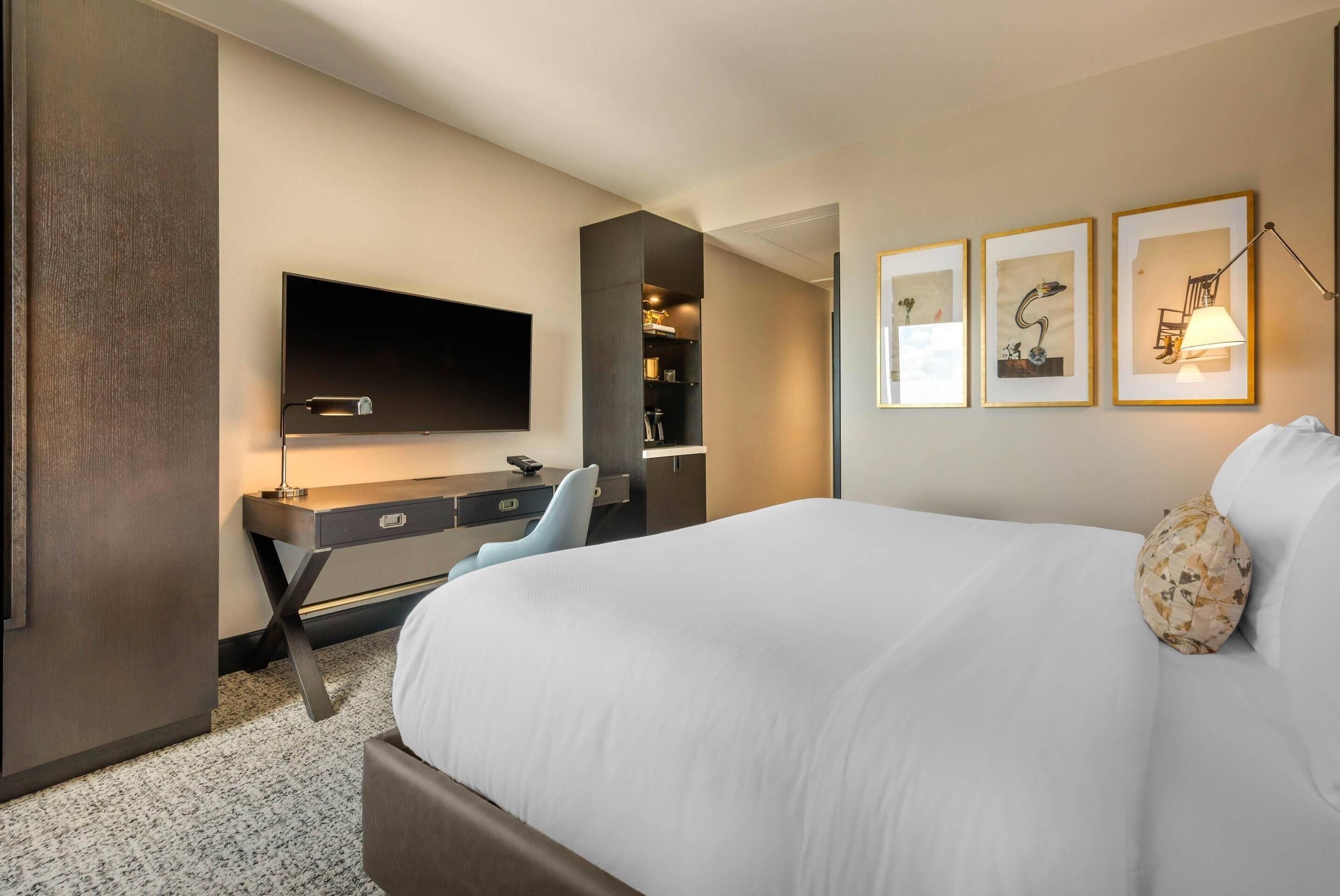 Quarto de hotel moderno com uma cama grande, uma mesa de trabalho com televisão de tela plana, estante embutida e obras de arte emolduradas na parede.