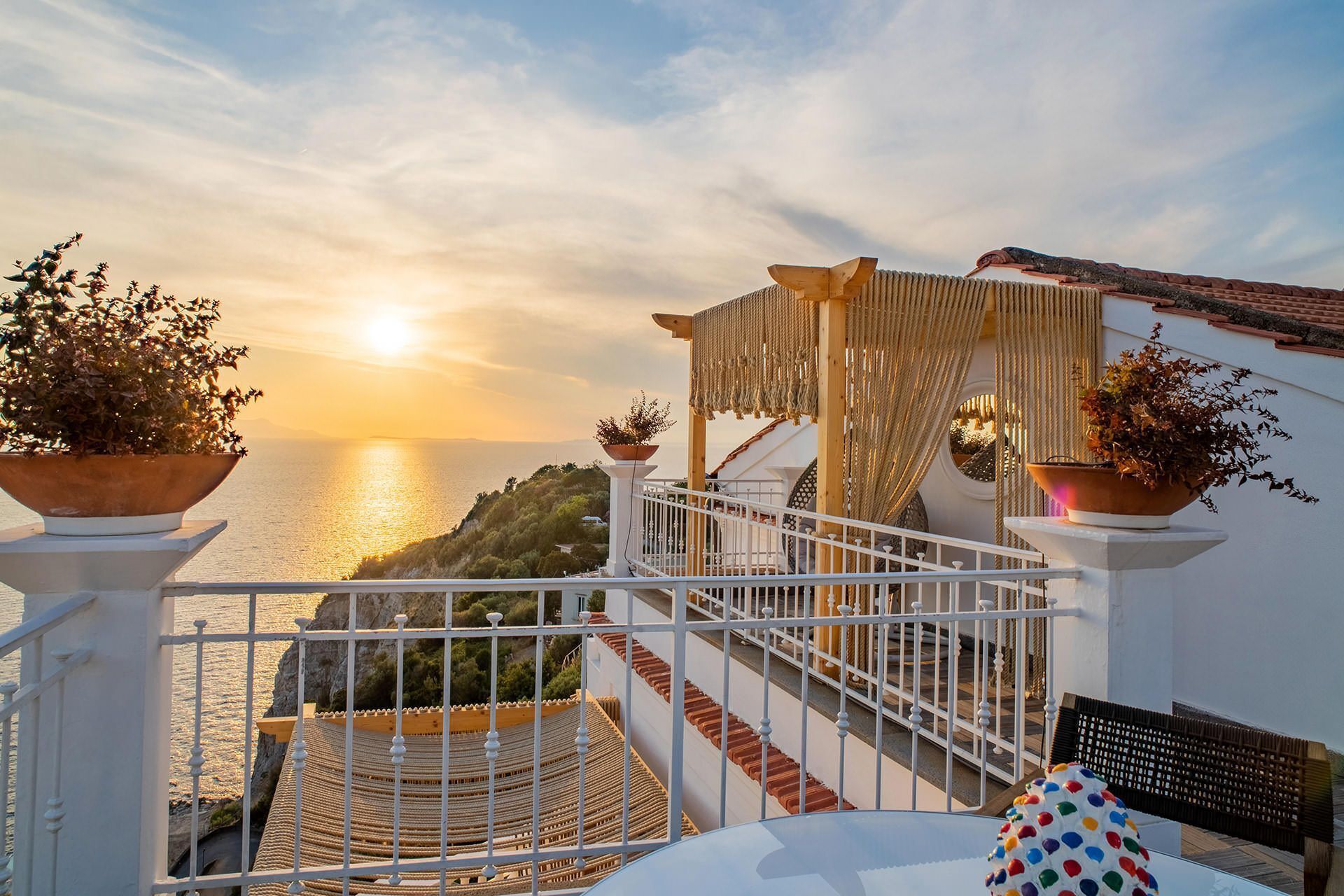 A balcony with a view of the sea at sunset, featuring a railing, potted plants, and a seating area.