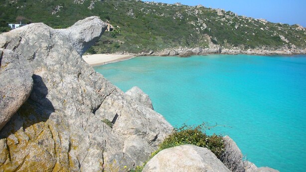 A coastal landscape with a clear turquoise sea, rocky outcrops, and a sandy beach.