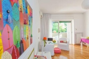 A room with a colorful wall art, a white chair, a small table with a laptop, and a view of greenery outside.