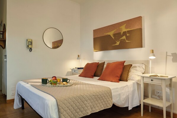 Una cama bien hecha con colcha, una mesita de noche con lámpara y un espejo montado en la pared.