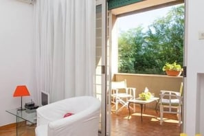 A room with a white bed, a glass table with a red lamp, and a balcony with chairs and a potted plant.