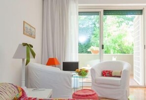 Ein Zimmer mit Bett, Stuhl, Tisch, Lampe und einem Fenster mit Blick auf Grünflächen.