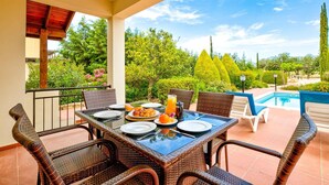 Een terras met een ontbijttafel, omringd door rieten stoelen, een zwembad en verzorgde begroeiing.
