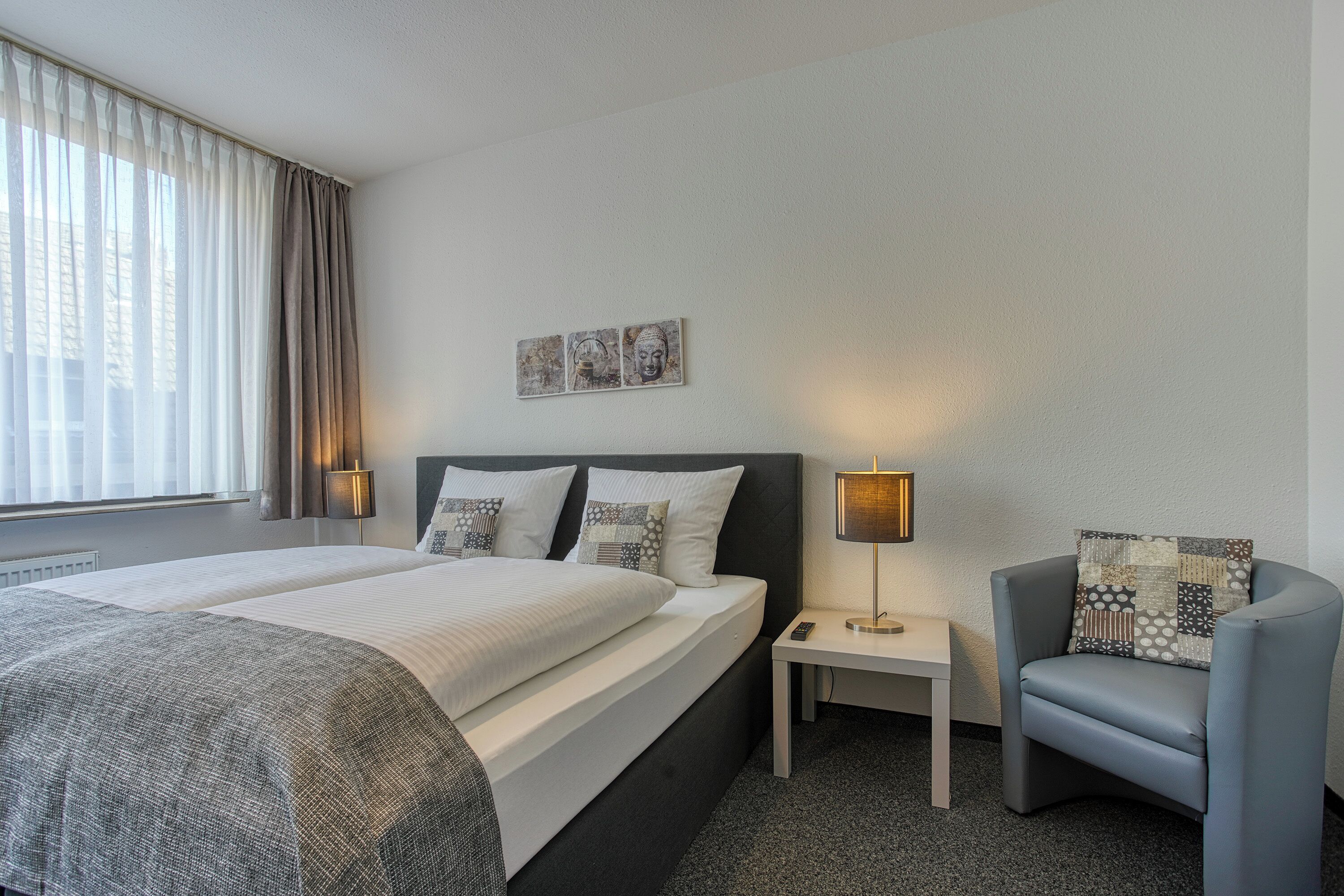 A hotel room with a large bed, a bedside table with a lamp, a chair, and a wall-mounted artwork.