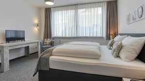 Ein modernes Hotelzimmer mit Bett, Schreibtisch mit Fernseher, Fenster mit Vorhängen und Wandbildern.