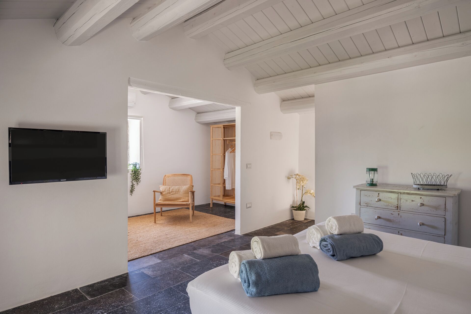 Una camera da letto moderna con una TV a schermo piatto, un letto con asciugamani arrotolati, una sedia di legno, un armadio e una piccola pianta.