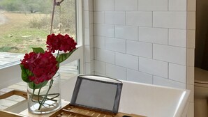A bathroom with white tiles, a window with a view of greenery, a wooden shelf with a tablet and a vase of flowers.